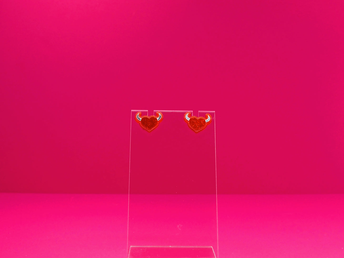 Devil horned heart earrings on a clear disay against a pink background.