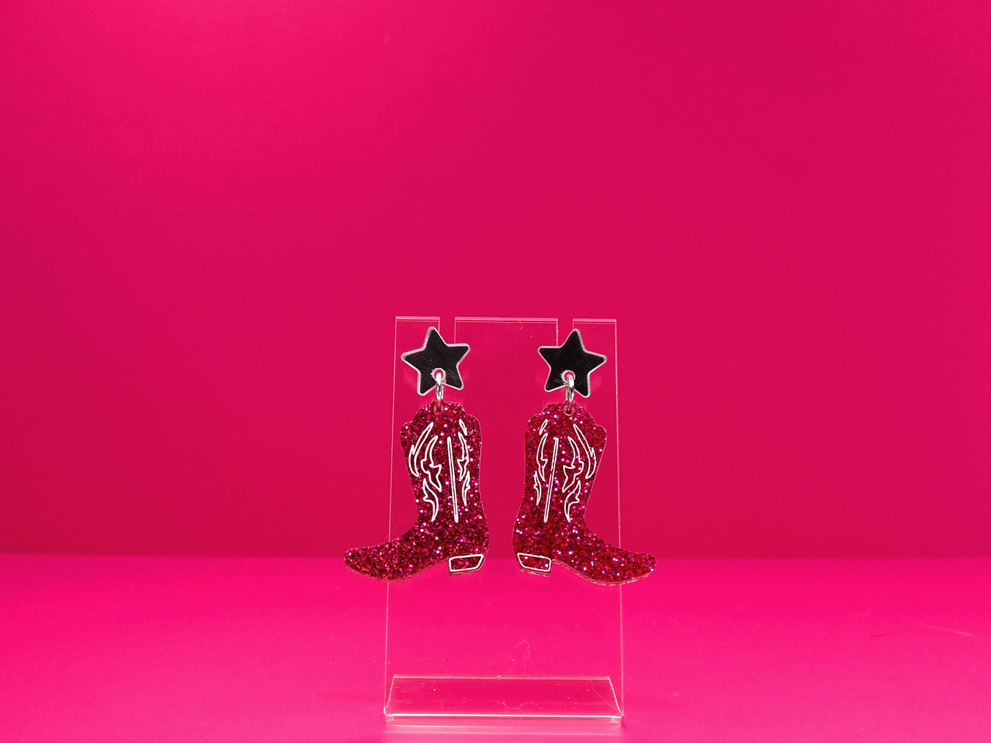 Pink cowboy boot earrings on a clear display against a pink background.