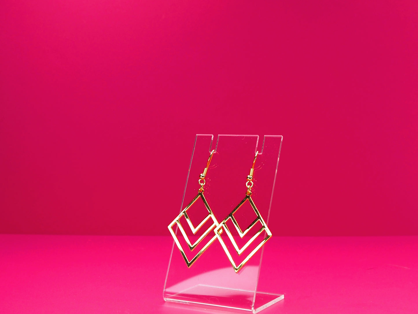 Gold geometric earrings on a clear display against a pink background.