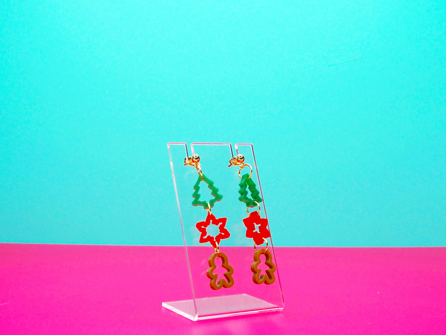 Christmas cookie cutter earrings on a clear display against a pink background.
