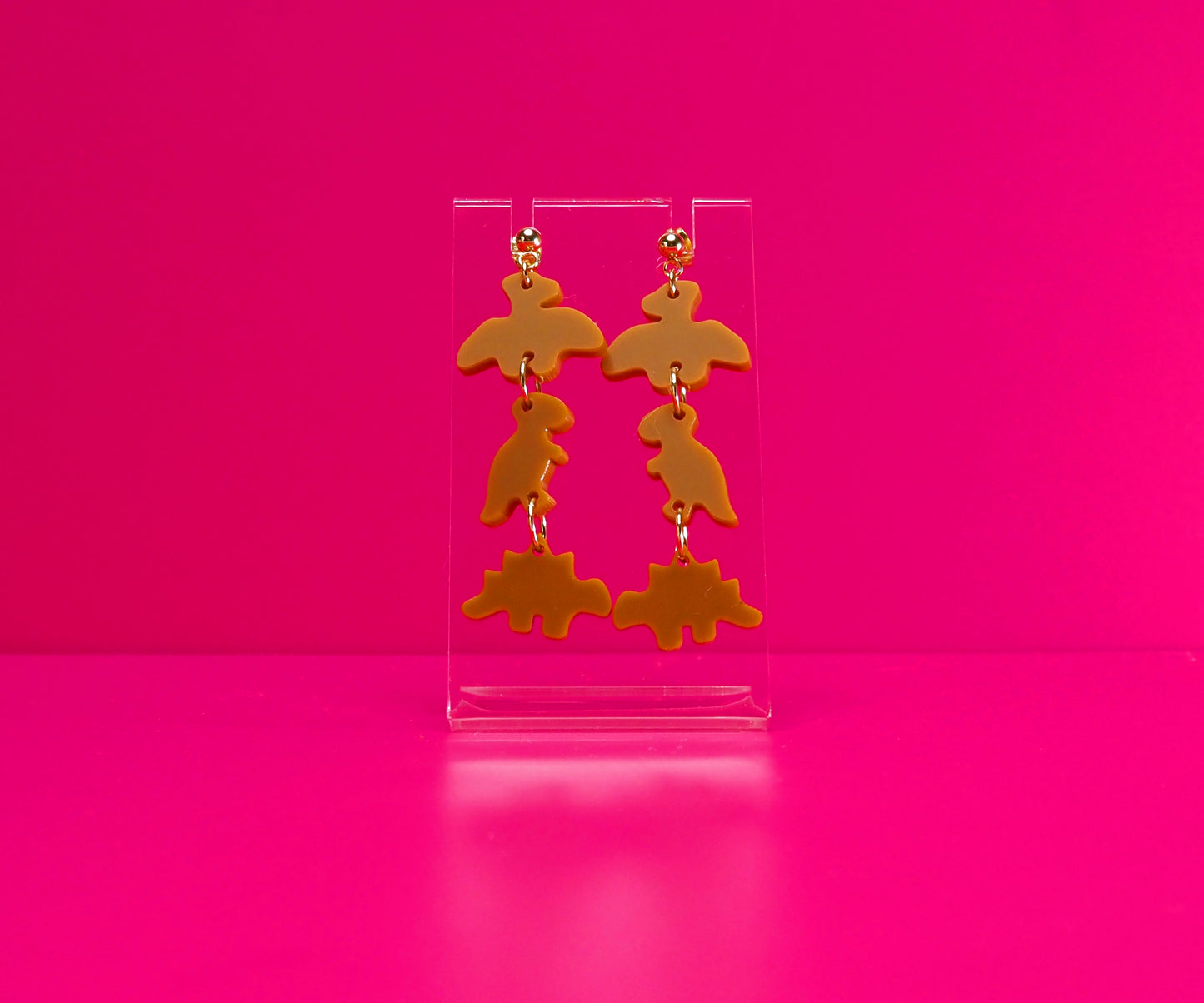 Dinosaur nugget earrings on a clear stand against a pink background.