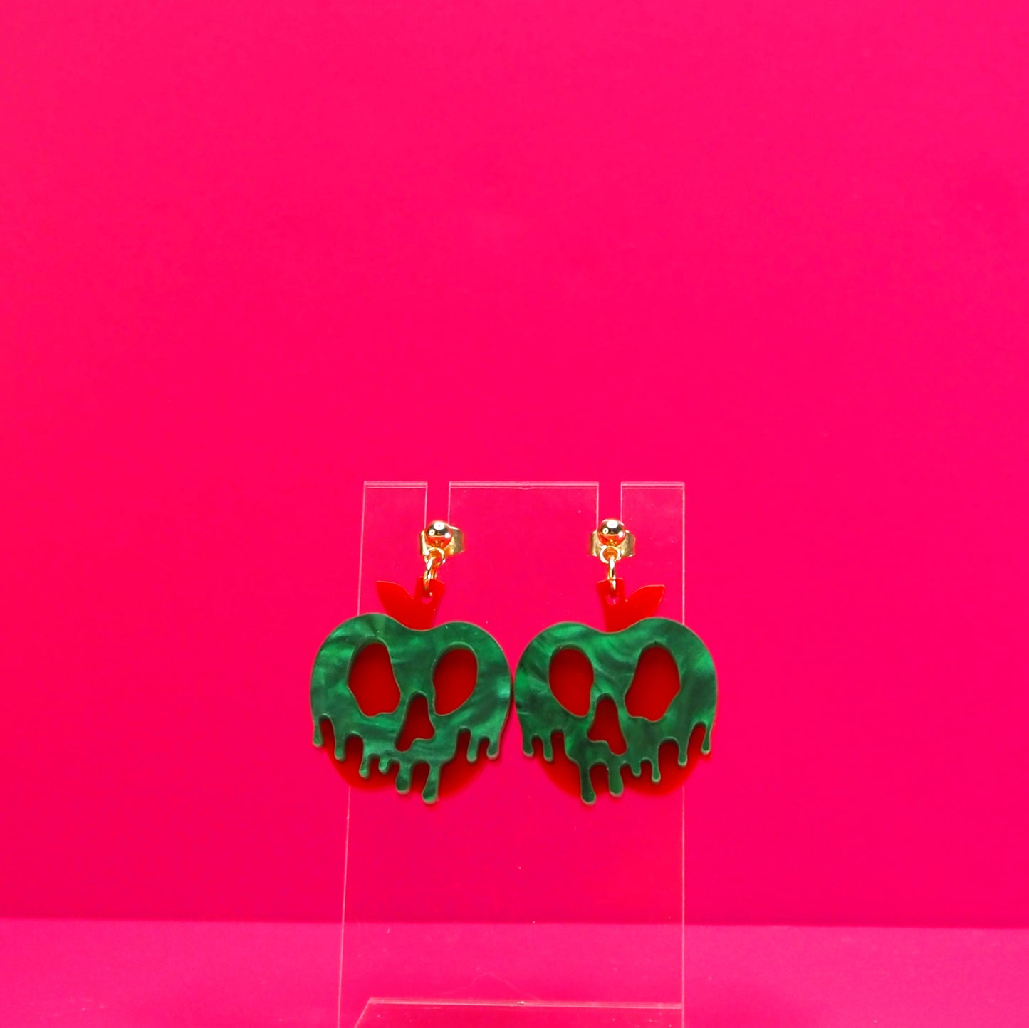 Red apple with green pearl goo earrings on a clear display against a pink background.