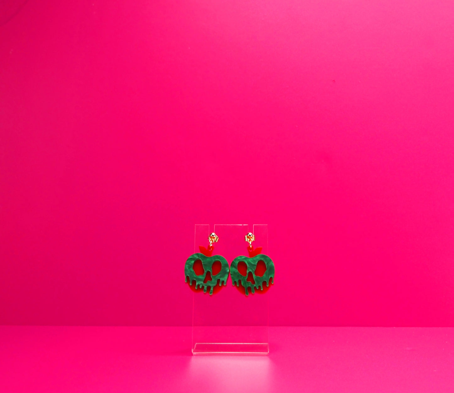 Red apple with green pearl goo earrings on a clear display against a pink background.