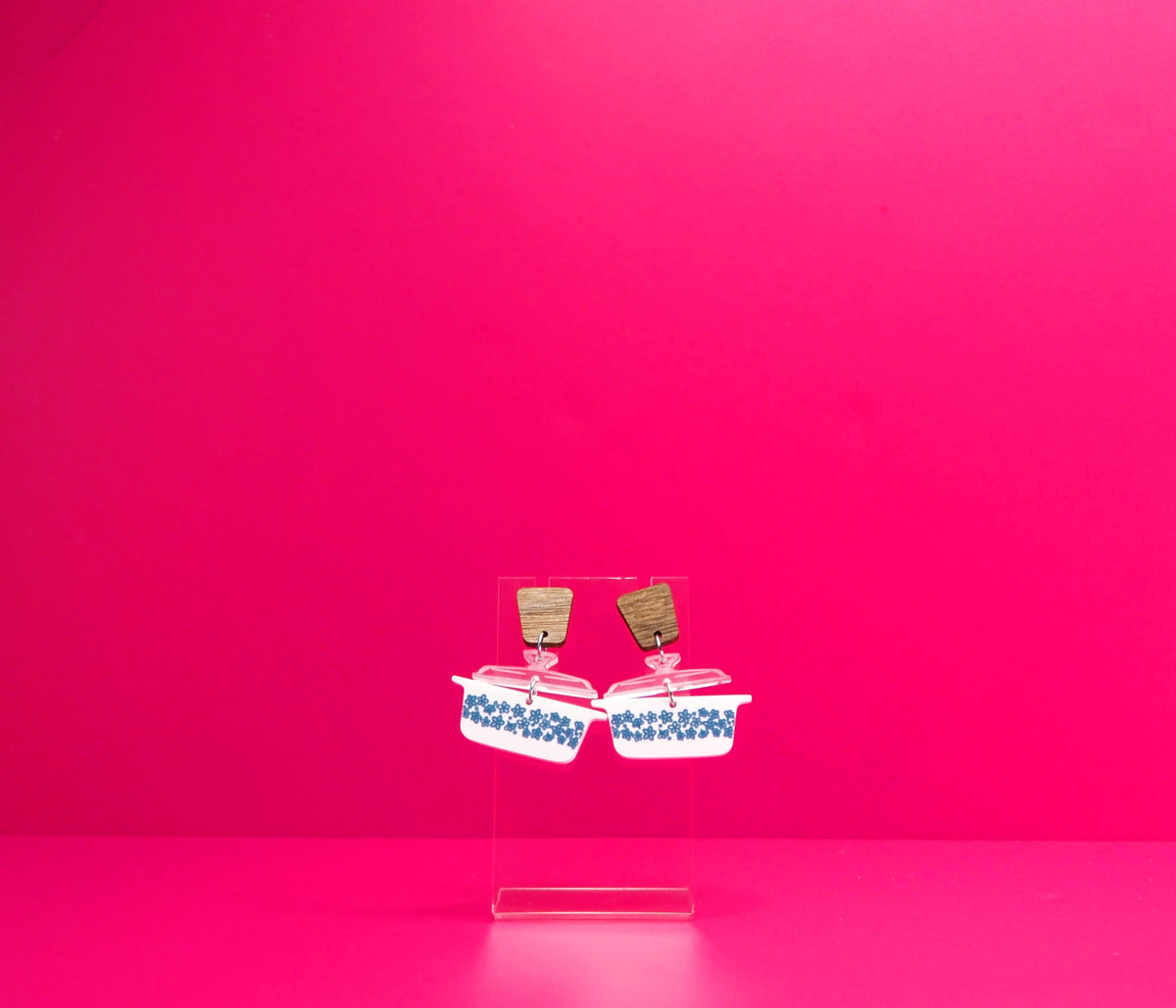 Vintage casserole dish earrings with wood accents on a clear display against a pink background.