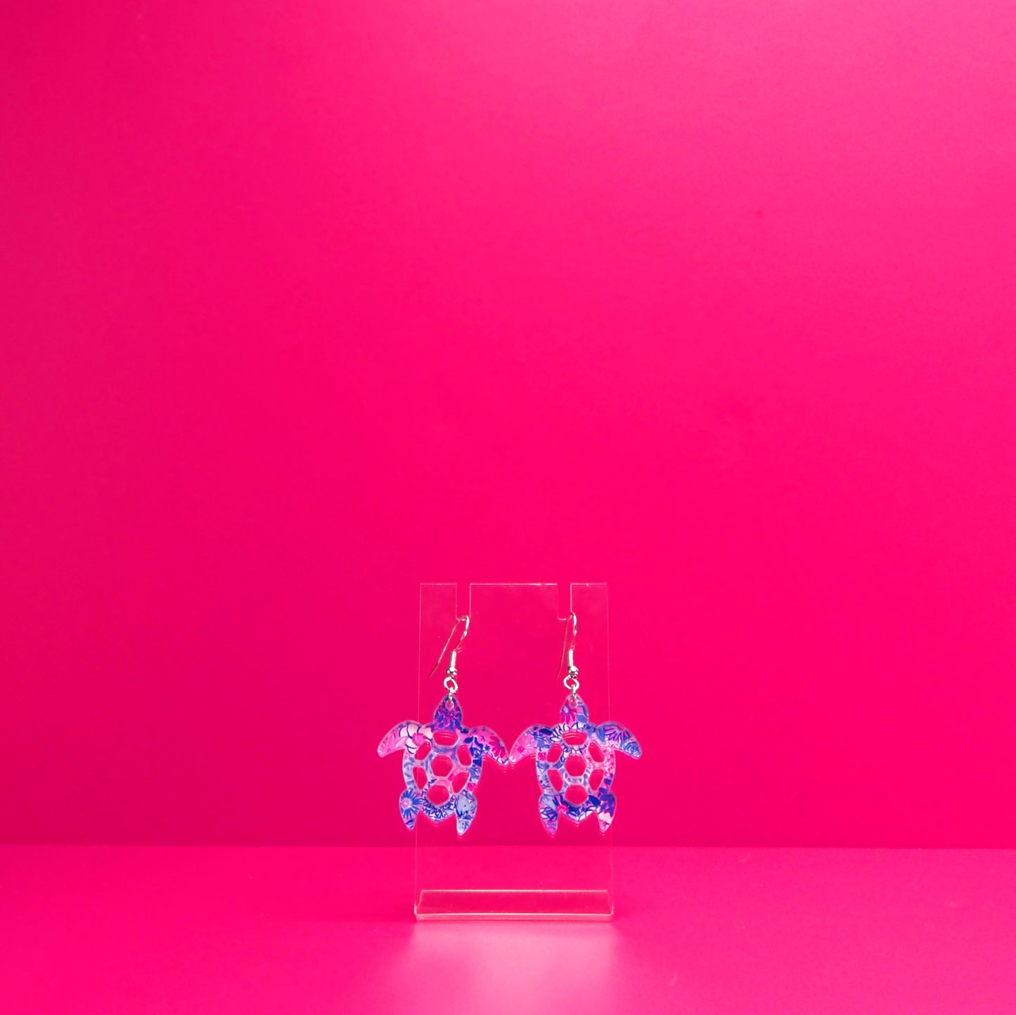 Purple blue, pink, white floral sea turtle earrings on a clear display against a pink background.