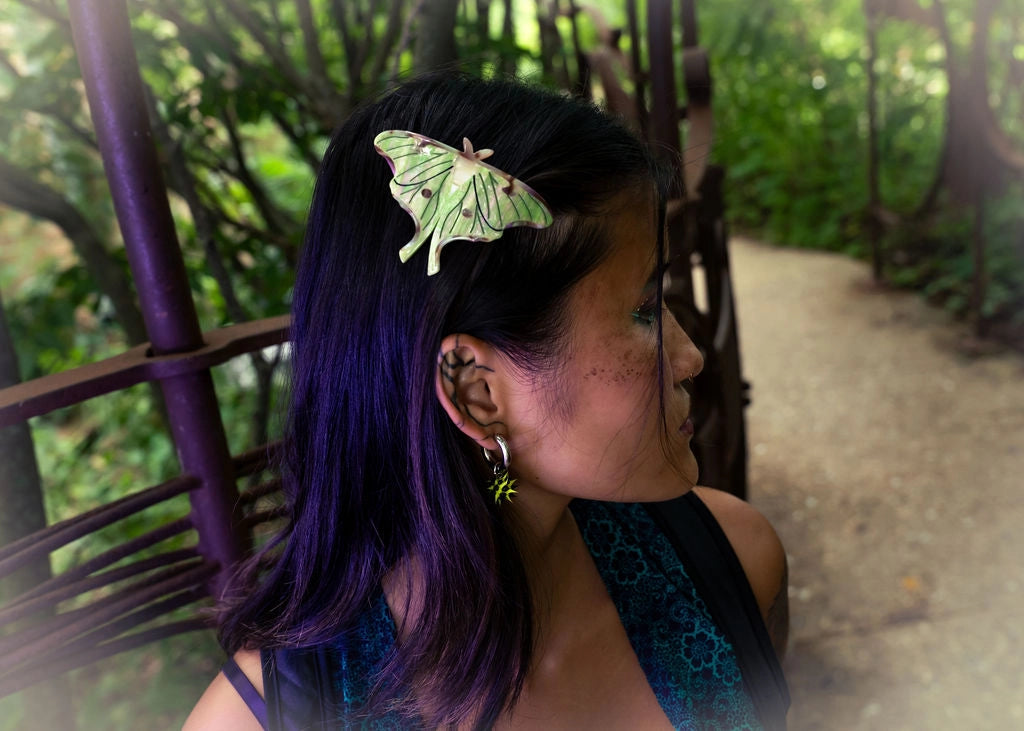 Multi-color haired model wearing green Luna Moth hair clip against a forest background.