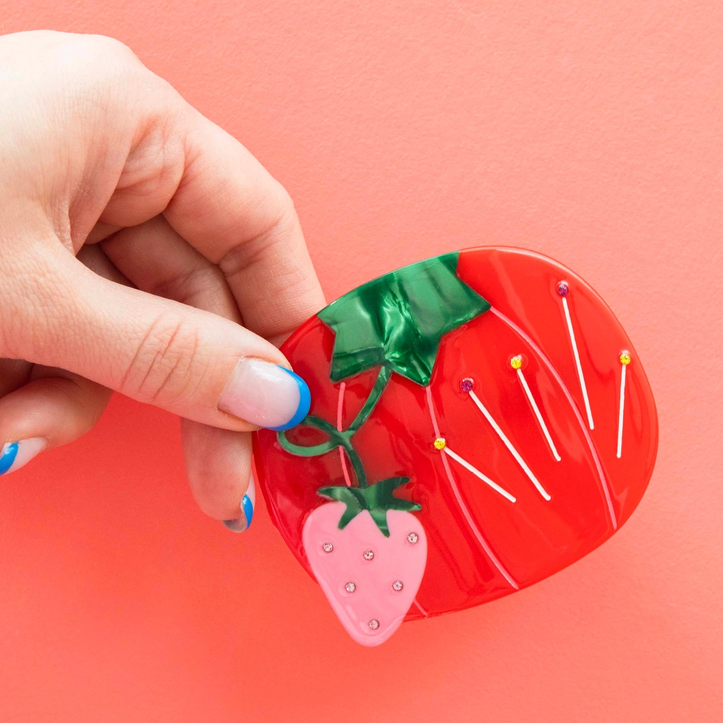 Model hand holding red tomato sewing pin hair barrette with a pink strawberry against a pink background.