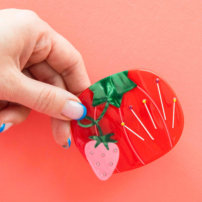 Model hand holding red tomato sewing pin hair barrette with a pink strawberry against a pink background.