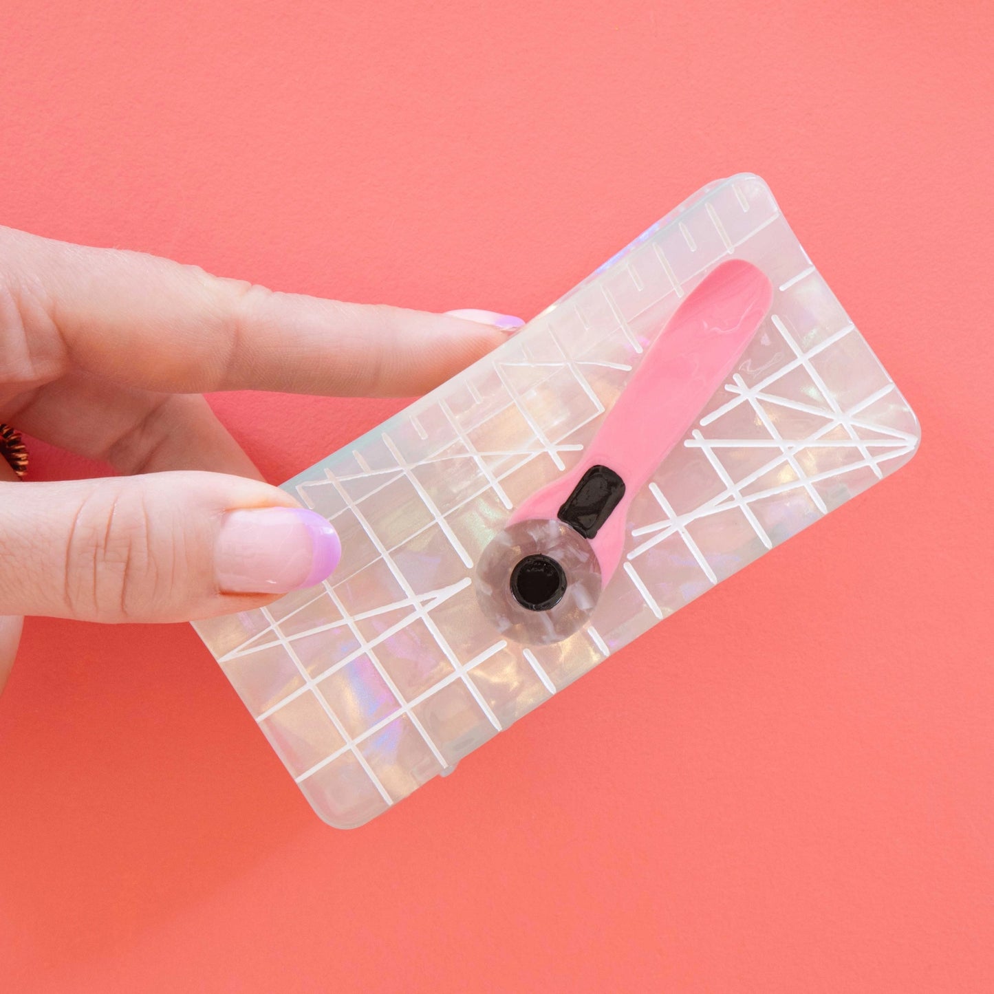 Model hand holding hair claw with iridescent cutting mat, and a pink rotary cutter against a pink background.