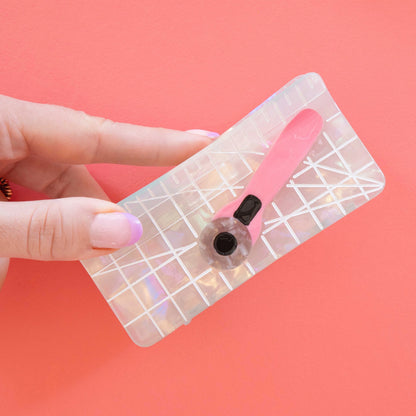 Model hand holding hair claw with iridescent cutting mat, and a pink rotary cutter against a pink background.