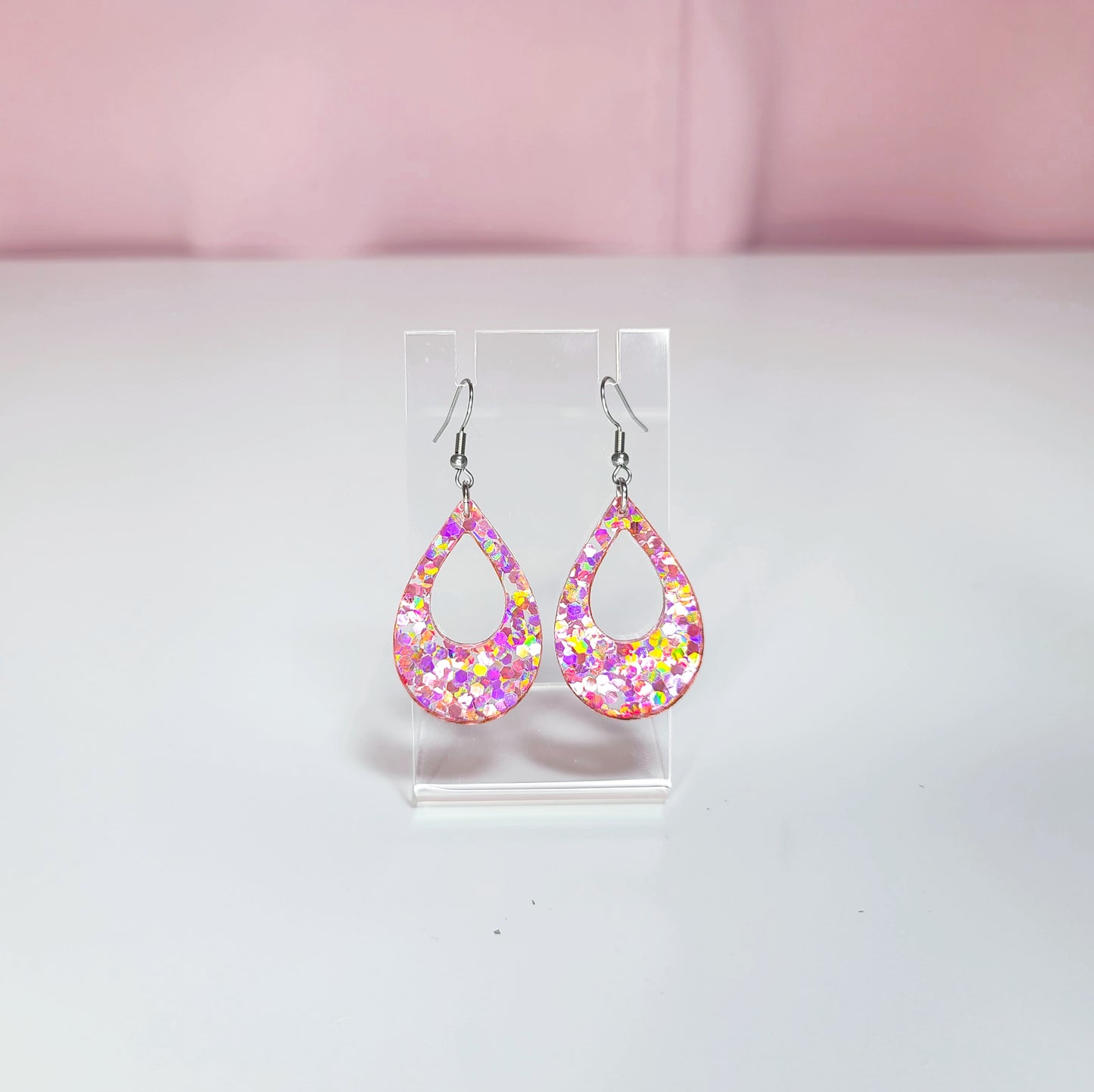 Earrings on a clear display against a white and pink background. Glitter in earrings give a yellow, purple, pink, silver color catch in the light