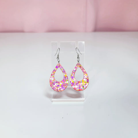 Earrings on a clear display against a white and pink background. Glitter in earrings give a yellow, purple, pink, silver color catch in the light