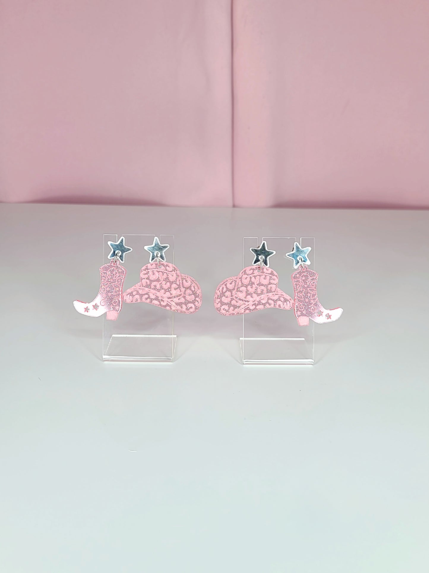 A pair of dangle earrings featuring pink cowboy boots with intricate designs, attached to mirror silver star studs. The earrings have a playful and retro aesthetic with a western theme. They are displayed on clear acrylic stands against a soft pink background.