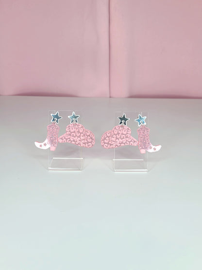 A pair of dangle earrings featuring pink cowboy boots with intricate designs, attached to mirror silver star studs. The earrings have a playful and retro aesthetic with a western theme. They are displayed on clear acrylic stands against a soft pink background.