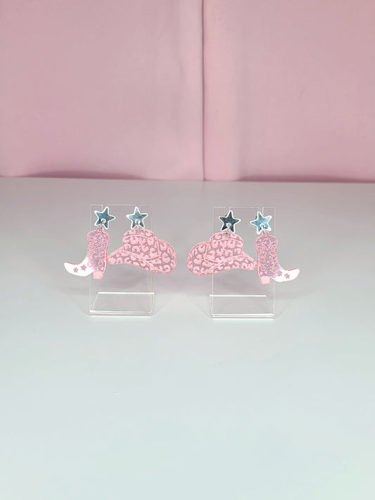 A pair of dangle earrings featuring pink cowboy boots with intricate designs, attached to mirror silver star studs. The earrings have a playful and retro aesthetic with a western theme. They are displayed on clear acrylic stands against a soft pink background.