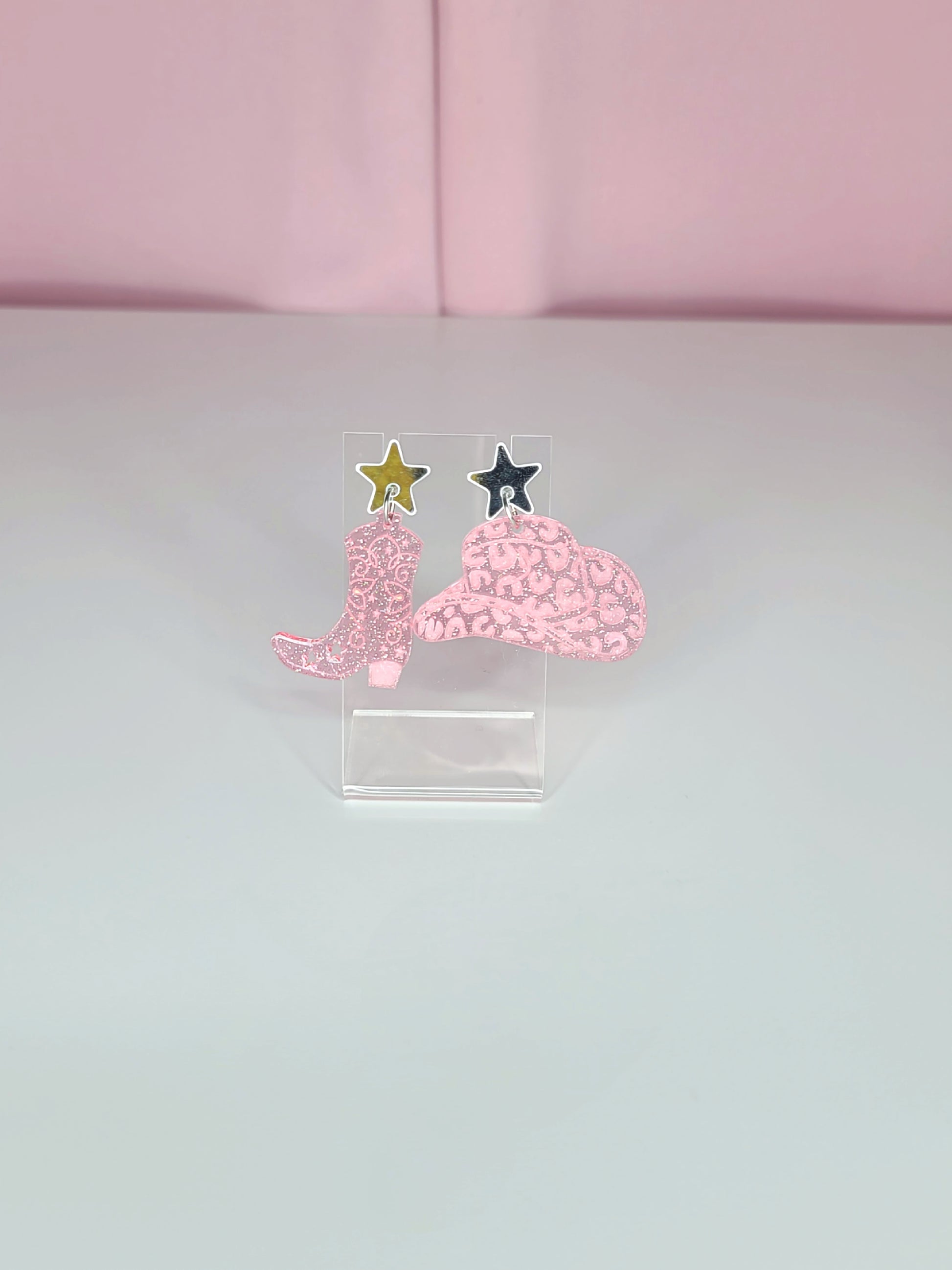 A pair of dangle earrings featuring pink cowboy boots with intricate designs, attached to mirror silver star studs. The earrings have a playful and retro aesthetic with a western theme. They are displayed on clear acrylic stands against a soft pink background.