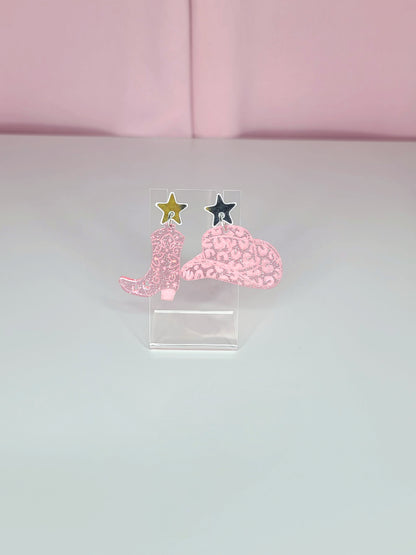 A pair of dangle earrings featuring pink cowboy boots with intricate designs, attached to mirror silver star studs. The earrings have a playful and retro aesthetic with a western theme. They are displayed on clear acrylic stands against a soft pink background.