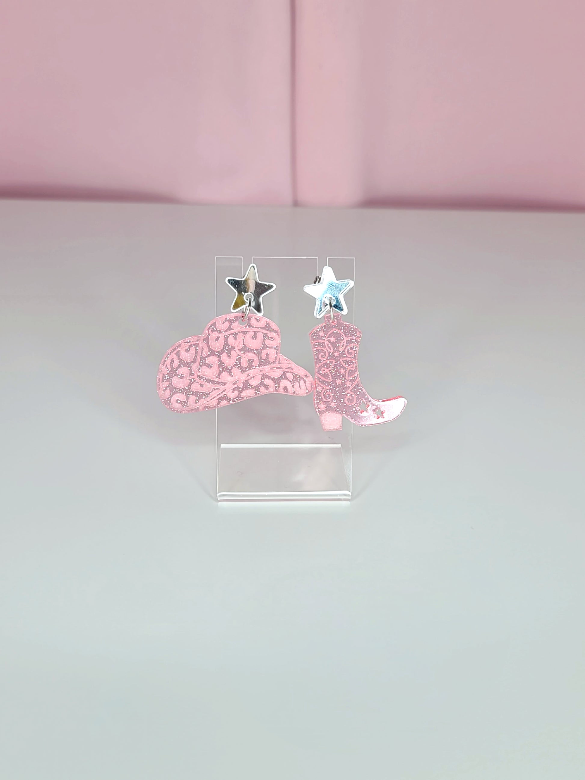 A pair of dangle earrings featuring pink cowboy boots with intricate designs, attached to mirror silver star studs. The earrings have a playful and retro aesthetic with a western theme. They are displayed on clear acrylic stands against a soft pink background.