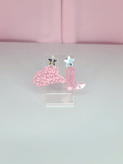 A pair of dangle earrings featuring pink cowboy boots with intricate designs, attached to mirror silver star studs. The earrings have a playful and retro aesthetic with a western theme. They are displayed on clear acrylic stands against a soft pink background.