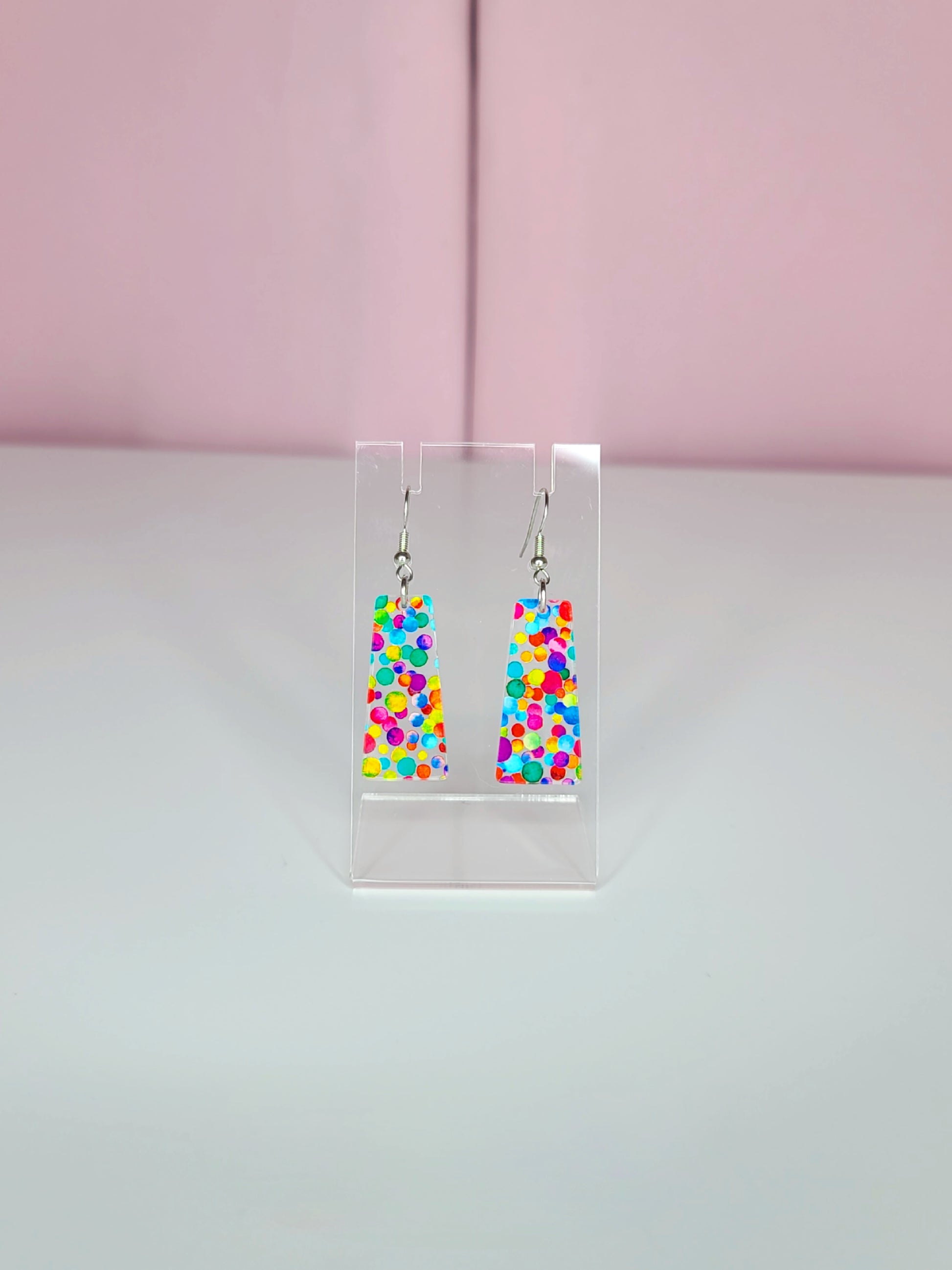 Earrings shown on a clear display against a pink and white background. Earrings feature rainbow dots that look like confetti