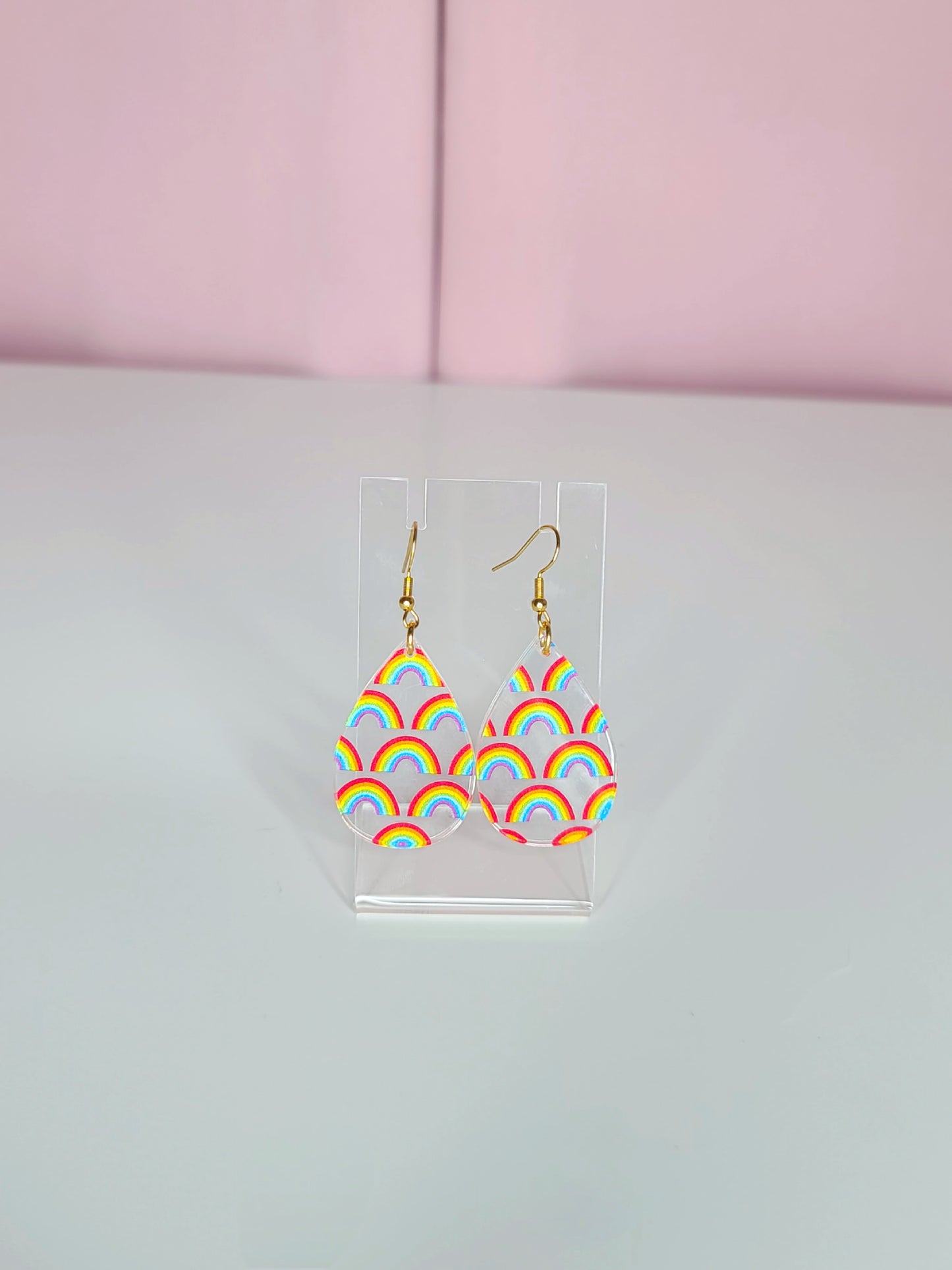 Clear teardrop-shaped earrings with delicate individual rainbow designs, displayed on a transparent stand against a soft white and pink background.