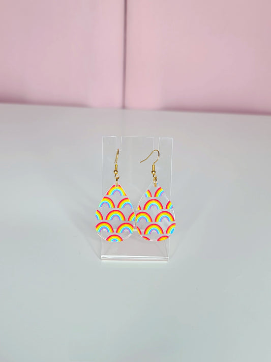 Clear teardrop-shaped earrings with delicate individual rainbow designs, displayed on a transparent stand against a soft white and pink background.