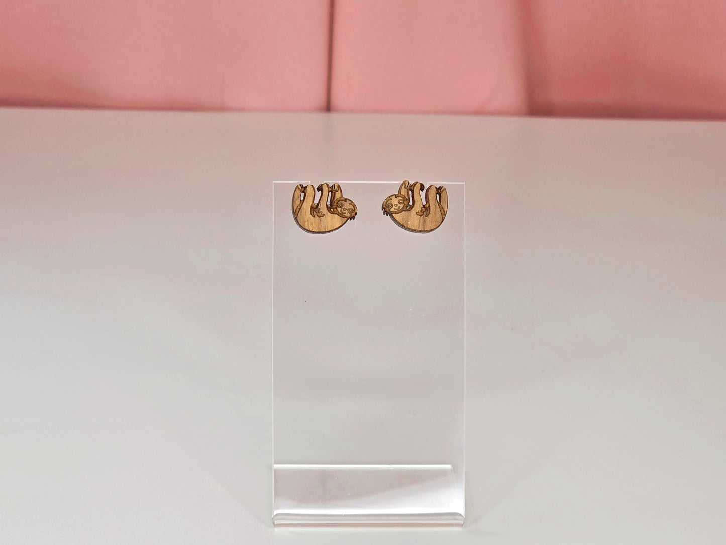 Dark brown wooden sloth earrings on a clear earring display against a pink and white background