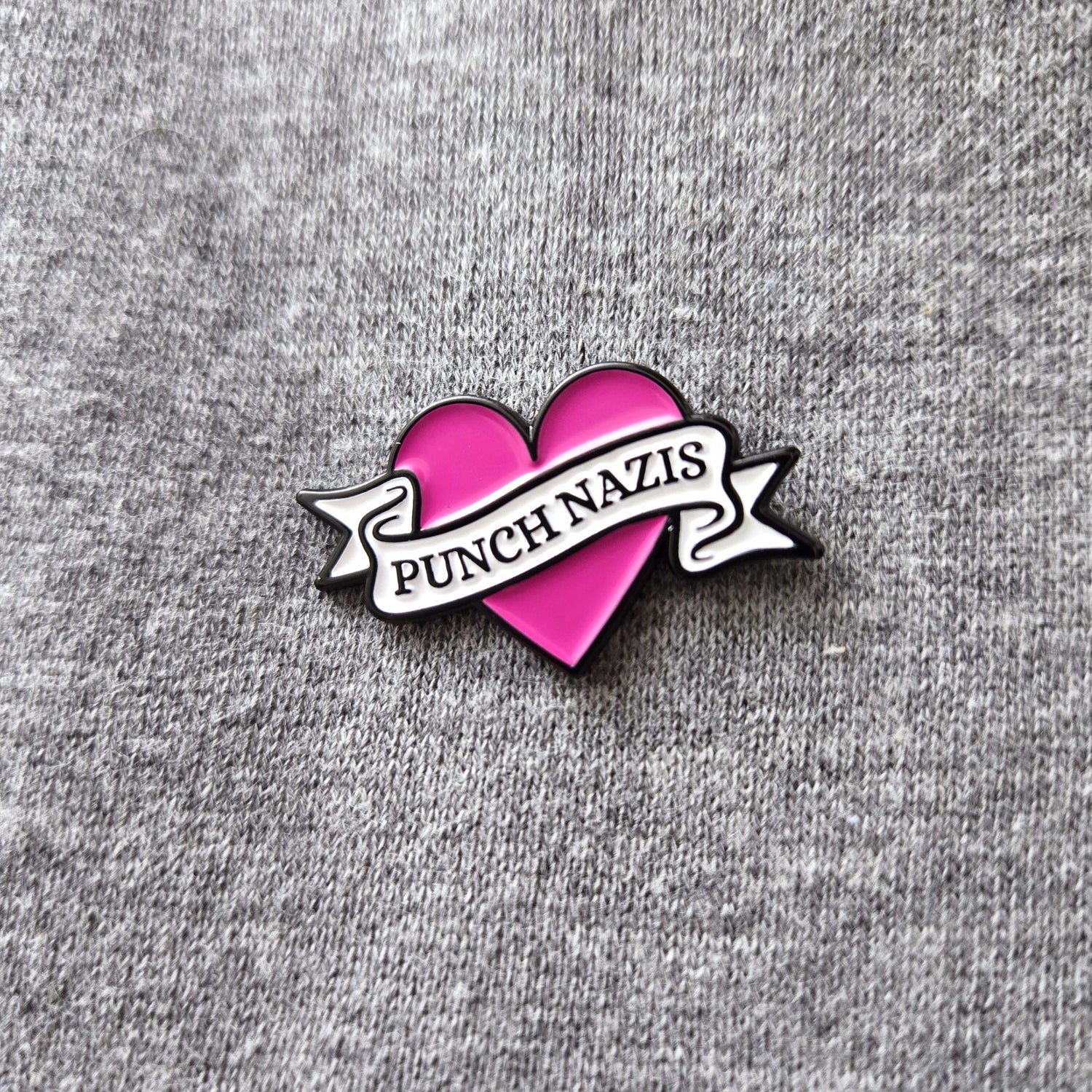 Pink heart with a white banner pin that states "Punch Nazis" against a grey background.