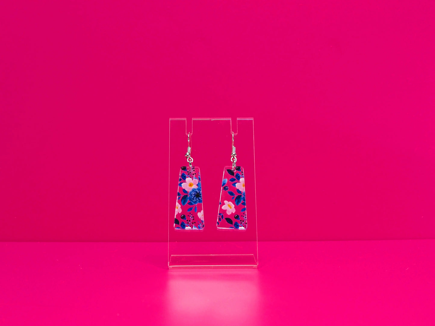 Blie and pink floral earrings on a clear stand against a pink background.