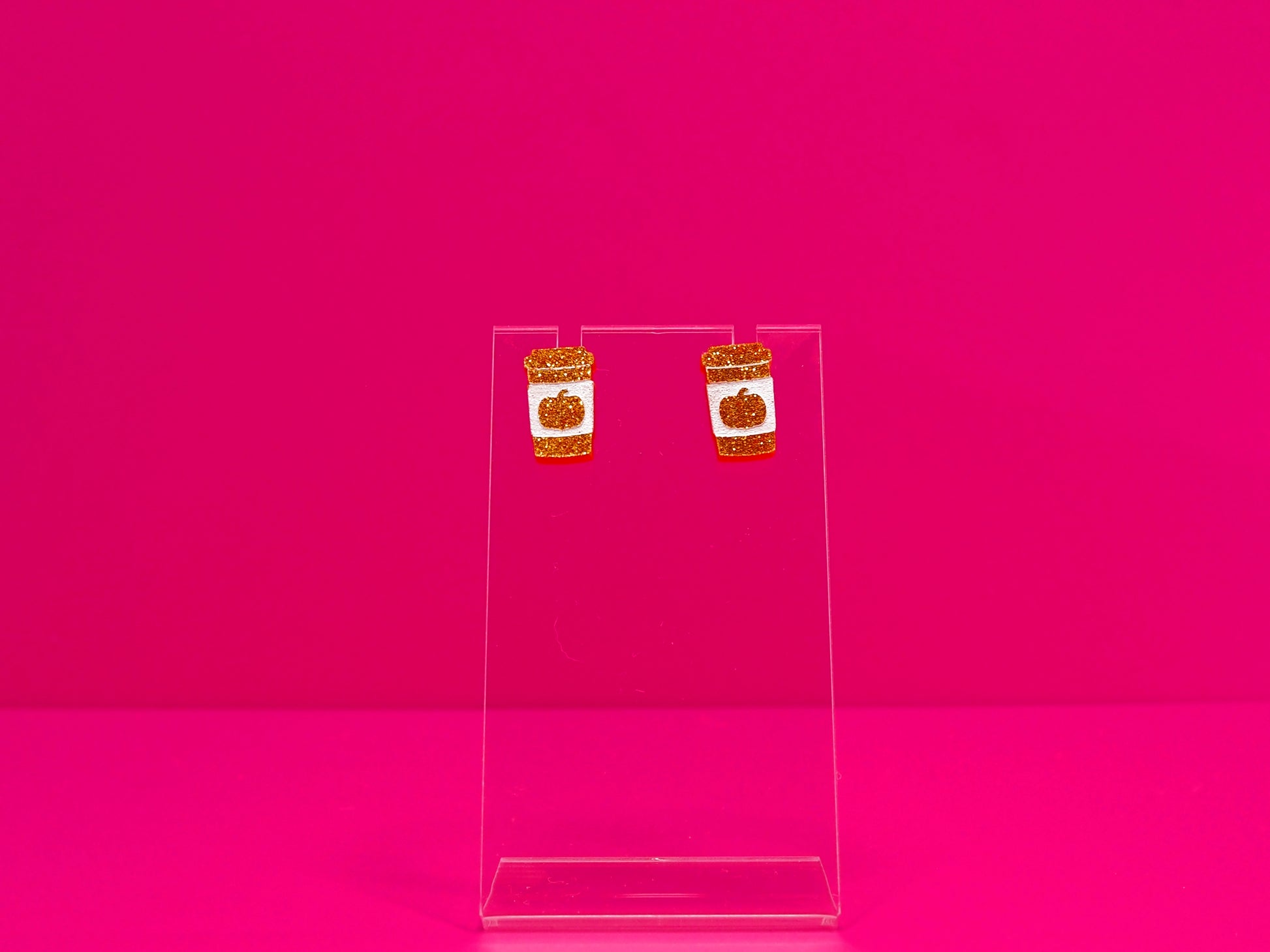 Orange flitter pumpkin spice cup earrings on a clear stand against a pink background.