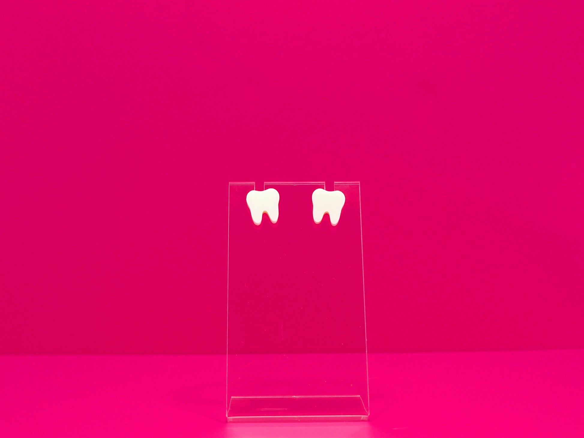 Pair of white tooth earrings on a clear display against a hot pink background.