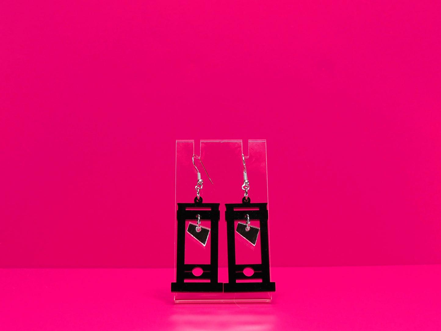 Black guillotine earrings on a clear display against a hot pink background.