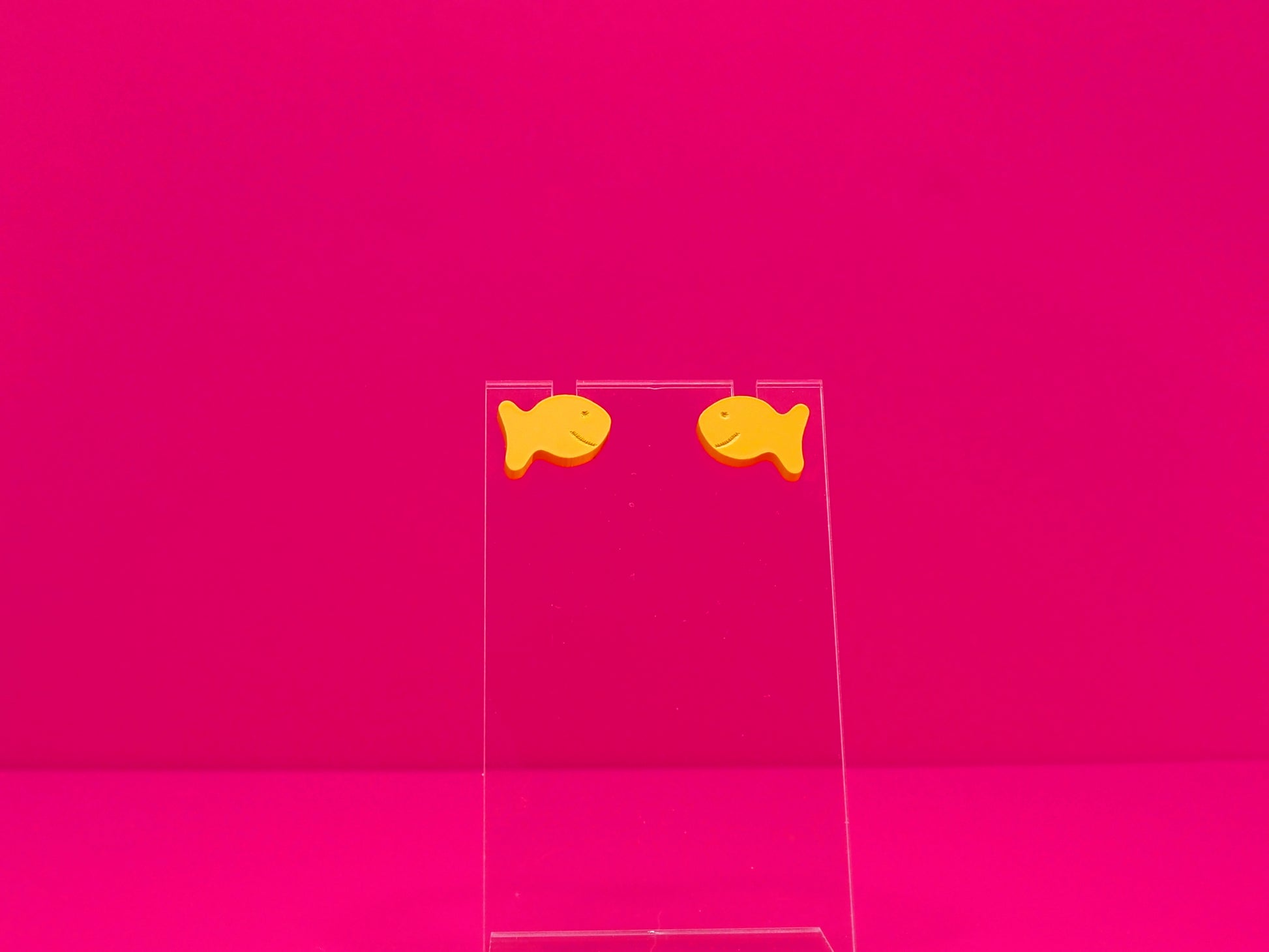 Two orange fish earrings on a clear display against a hot pink background.
