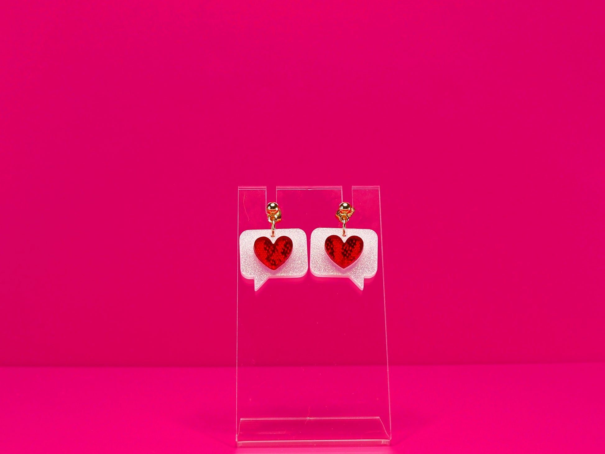 Red hearts on white text bubble earrings on a clear display against a pink background. Shown with gold studs option.