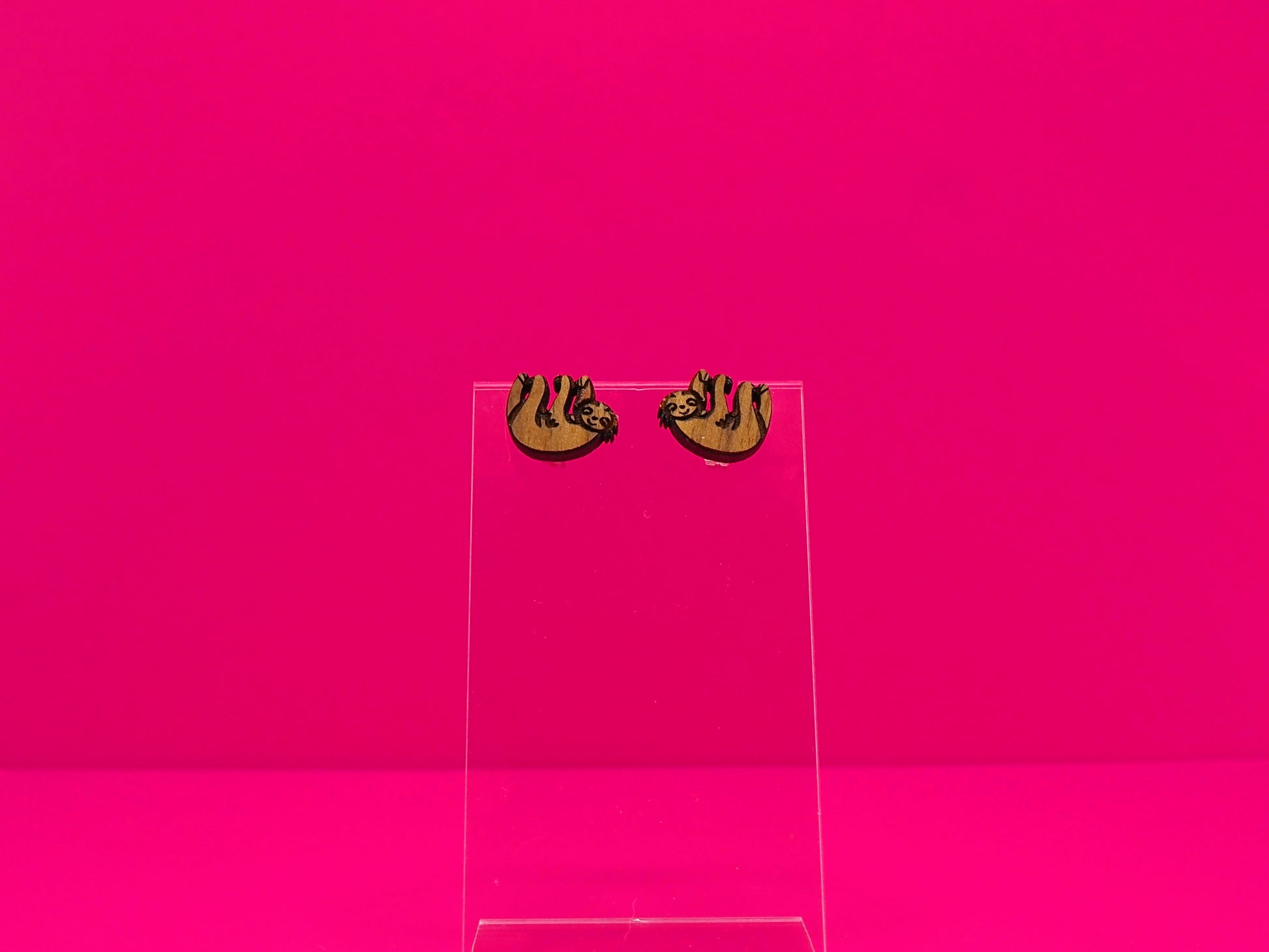 Pair of brown wood sloth earrings on a clear display against a hot pink background.