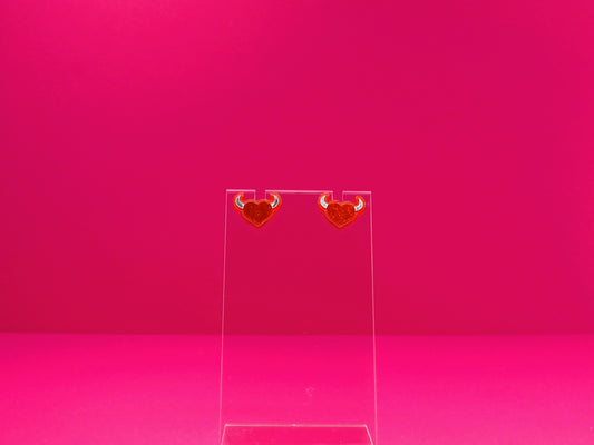 Devil horned heart earrings on a clear disay against a pink background.