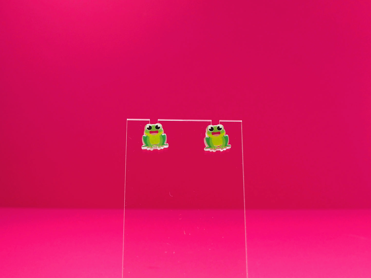 Frog earrings on a clear display against a pink background.