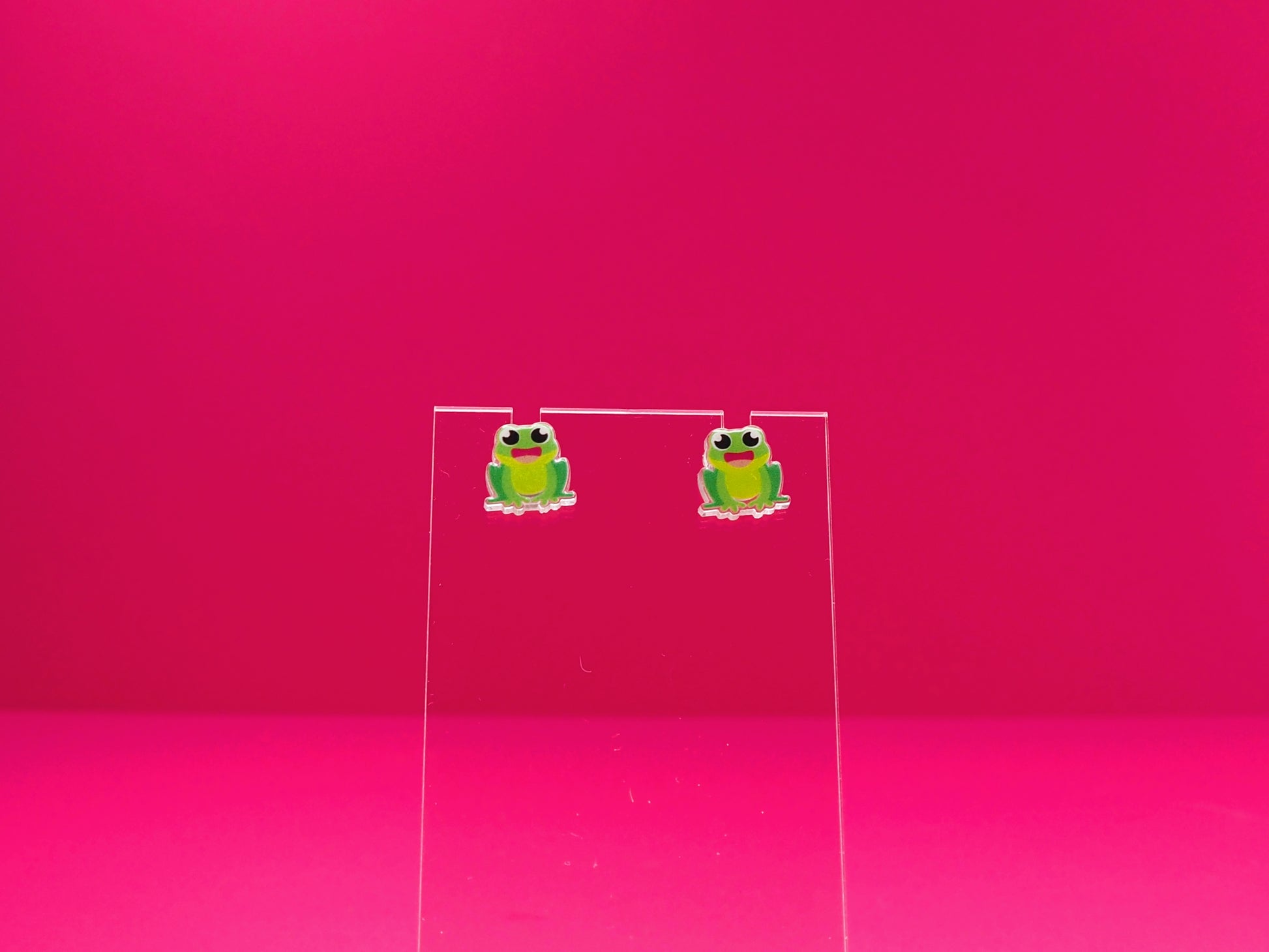 Frog earrings on a clear display against a pink background.