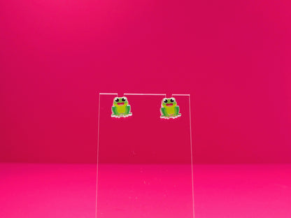 Frog earrings on a clear display against a pink background.