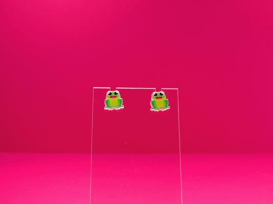 Frog earrings on a clear display against a pink background.