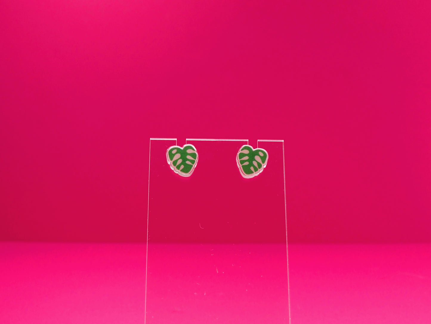 Monstera leaf earrings on a clear display against a pink background.