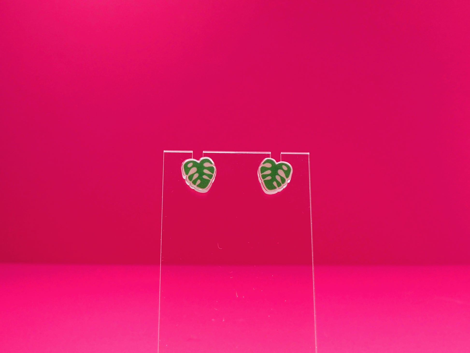 Monstera leaf earrings on a clear display against a pink background.