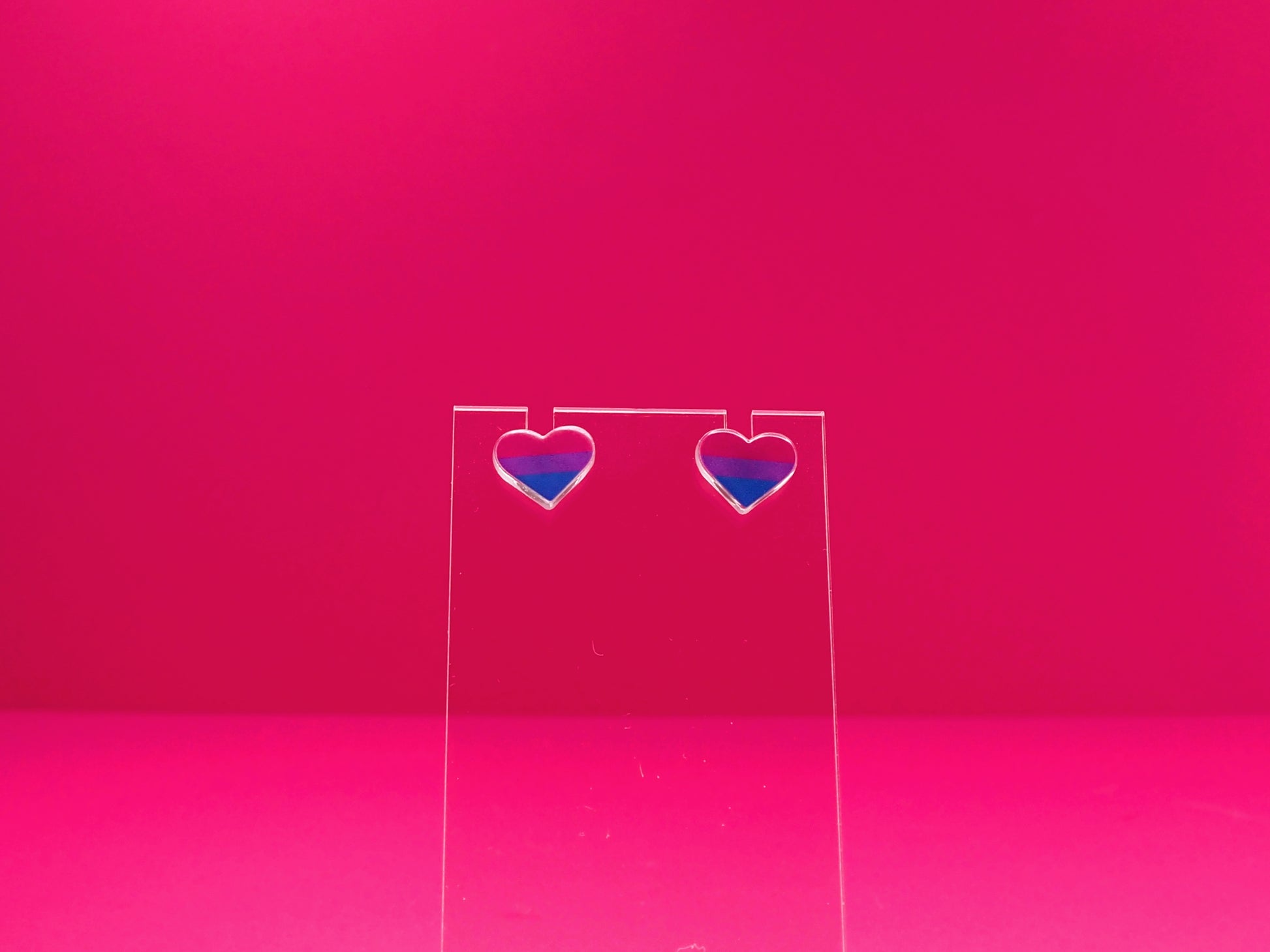 Bisexual pride heart earrings on a clear display against a pink background.