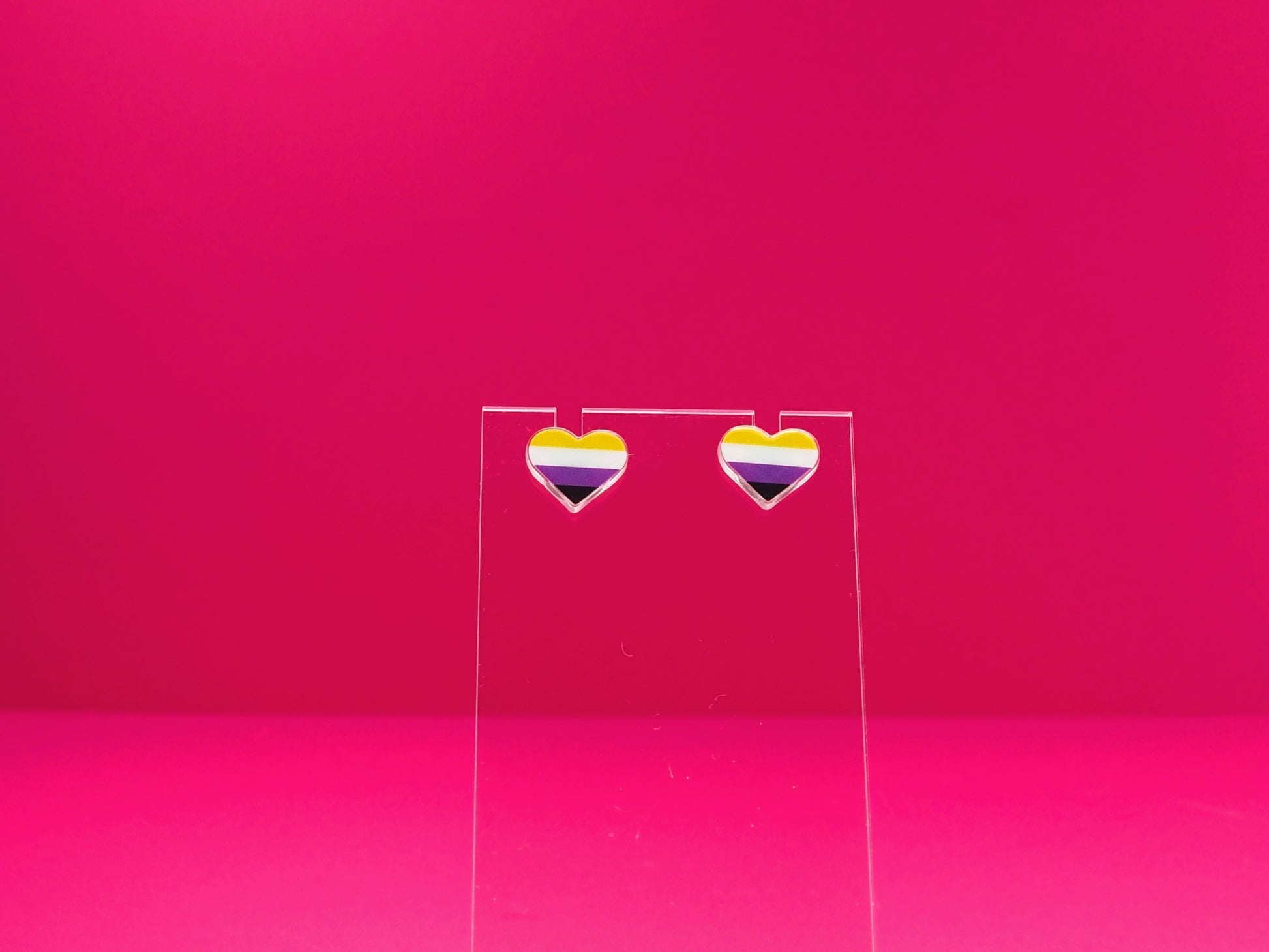 Non-binary pride heart earrings on a clear display against a pink background.