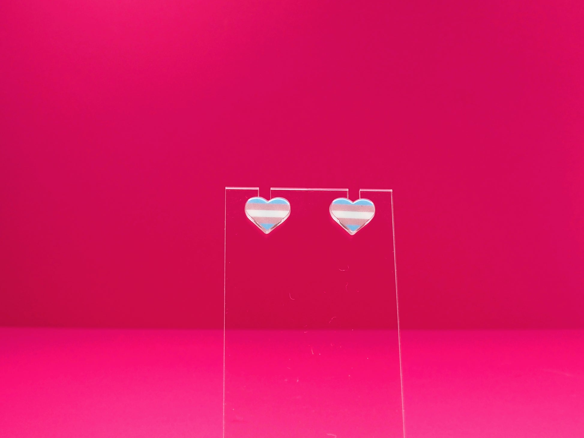 Transexual pride heart earrings on a clear display against a pink background.