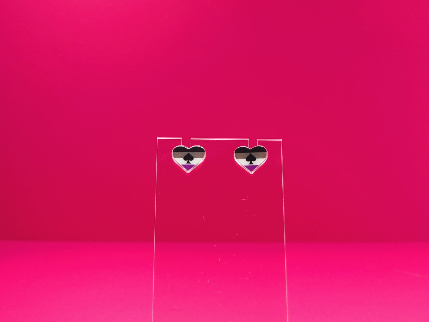 Ace pride heart earrings on a clear display against a pink background.