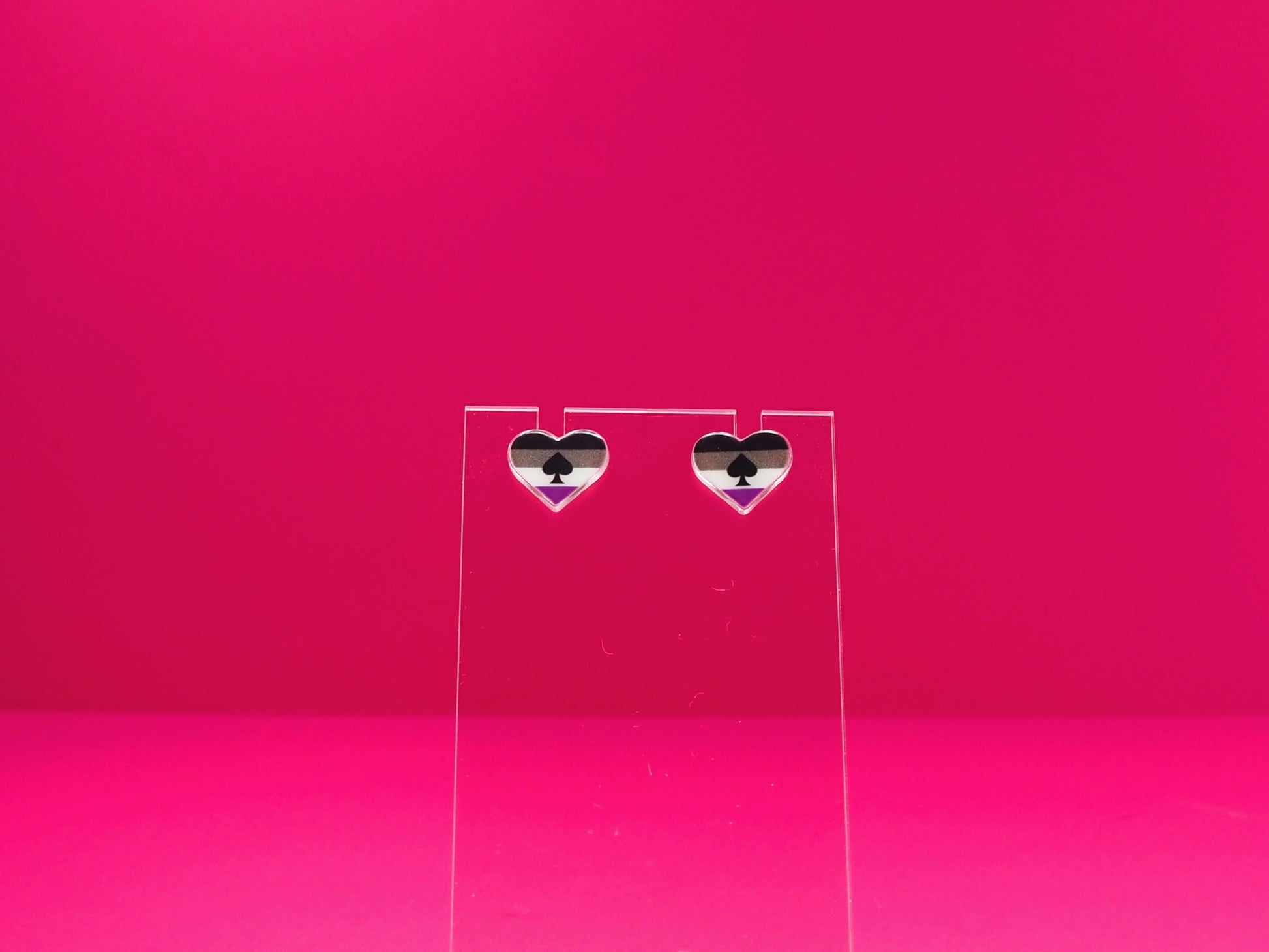 Ace pride heart earrings on a clear display against a pink background.