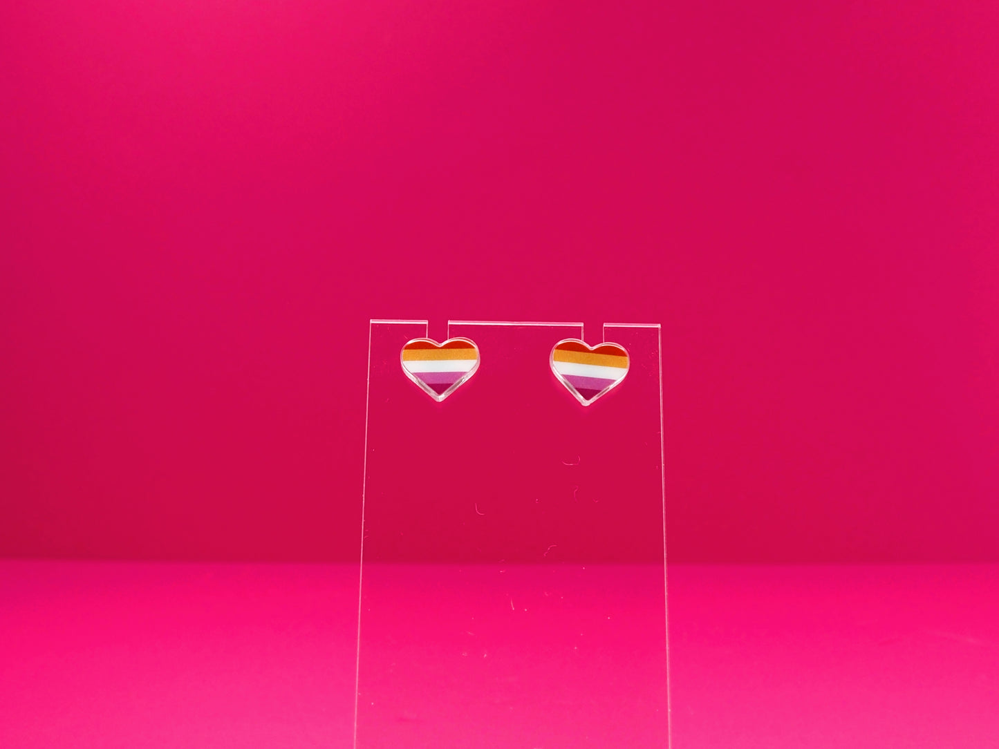 Lesbian pride heart earrings on a clear display against a pink background.