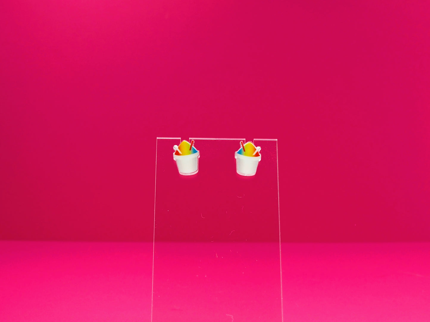Rainbow snoball earrings on a clear display against a pink background.