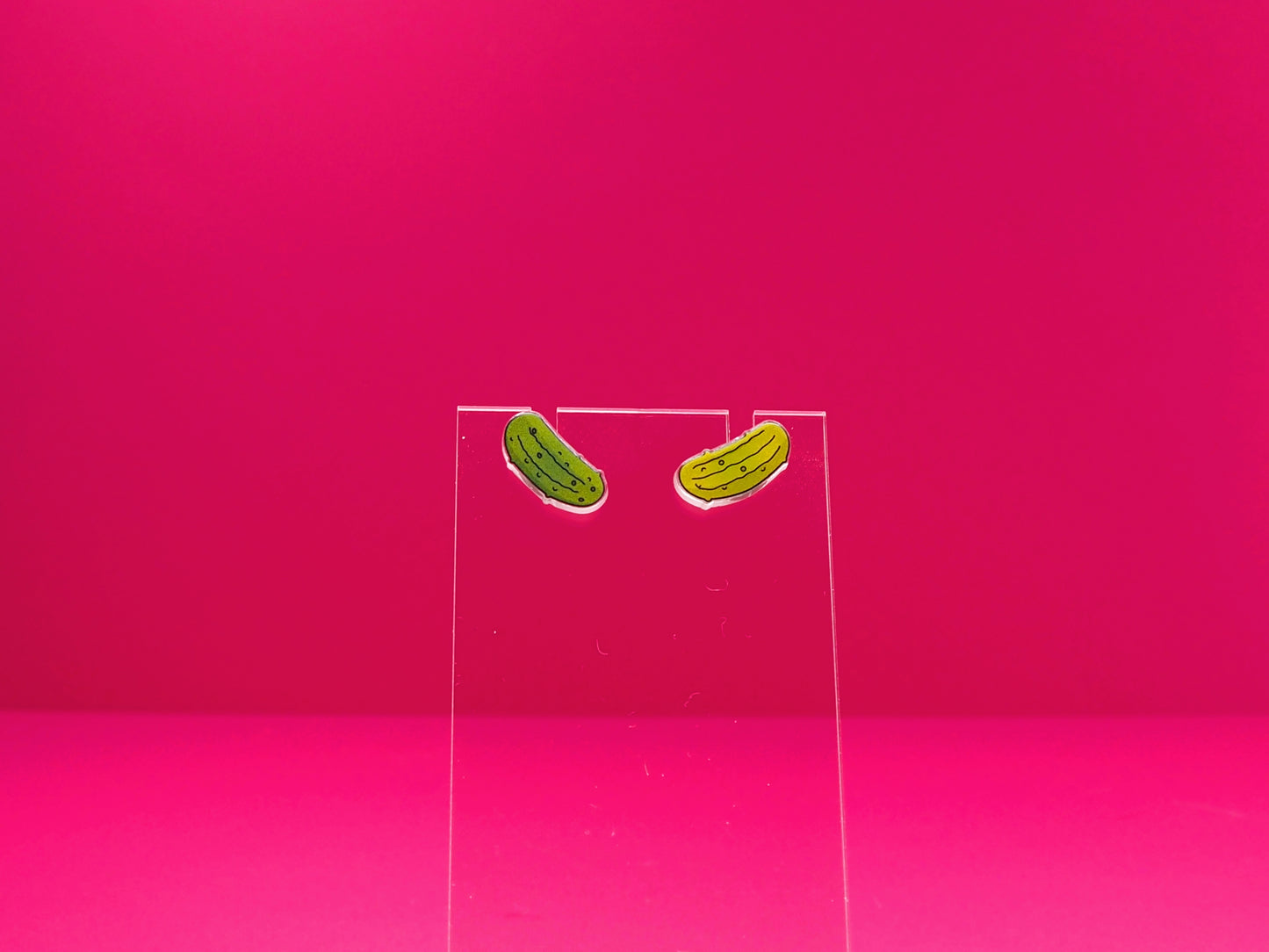Pickle earrings on a clear display against a pink background.
